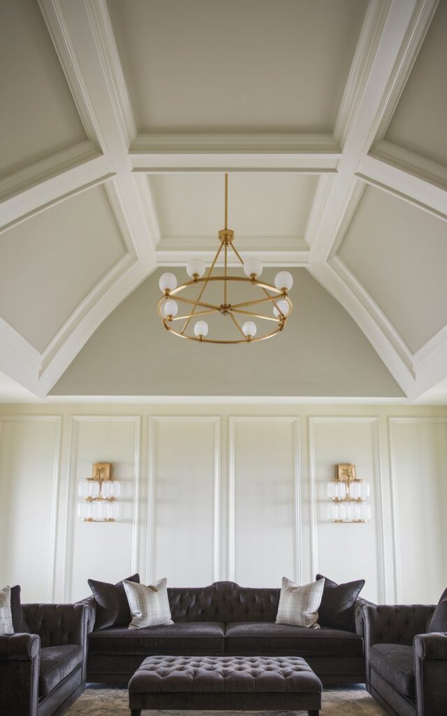 A photo of a sophisticated living room with a vaulted ceiling and a coffered ceiling design. The ceiling is painted in a soft white hue, adding depth and refinement to the room. Below the ceiling, there is a cozy sofa in dark charcoal fabric, providing a striking contrast. The sofa is complemented by a tufted ottoman and elegant gold-accented lighting fixtures. The overall aesthetic of the room blends modern comfort with timeless sophistication.