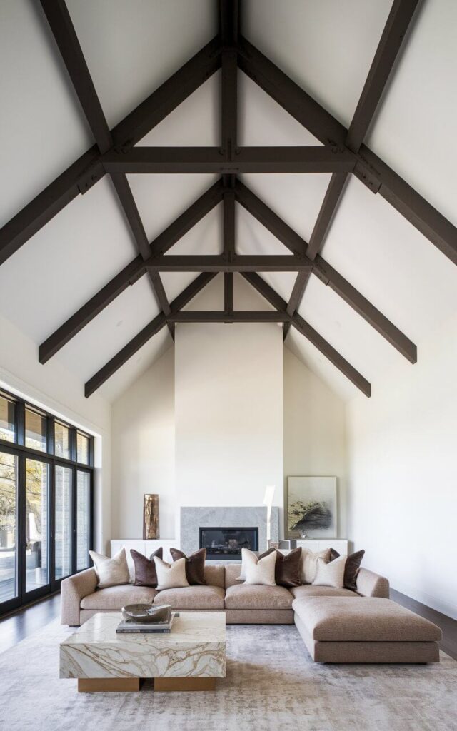 A captivating photo of a living room with a striking vaulted ceiling. The exposed beams are painted a deep espresso brown, creating a bold contrast against the crisp white ceiling. This contrast adds visual interest and enhances the room's architectural appeal. Below, a very cozy sofa in a soft oatmeal fabric pairs beautifully with a marble-top coffee table. Floor-to-ceiling windows let in natural light, balancing the darker ceiling elements.