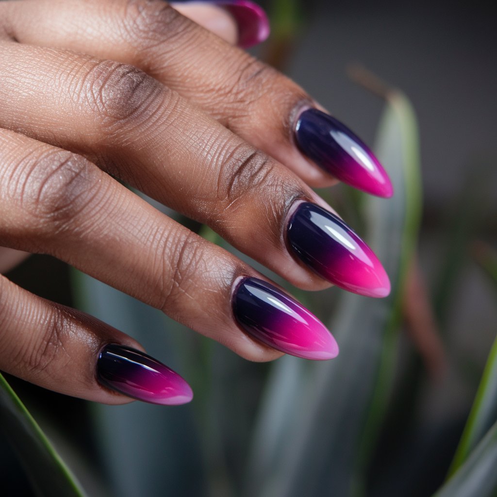 A close-up shot of a black woman's left hand with a dark purple-to-hot pink gradient manicure. The nails are almond-shaped and elongate the fingers. The skin is flawless, and the cuticles are well-groomed. The glossy finish of the nails reflects the natural background lighting. The background is blurred and composed of plants.