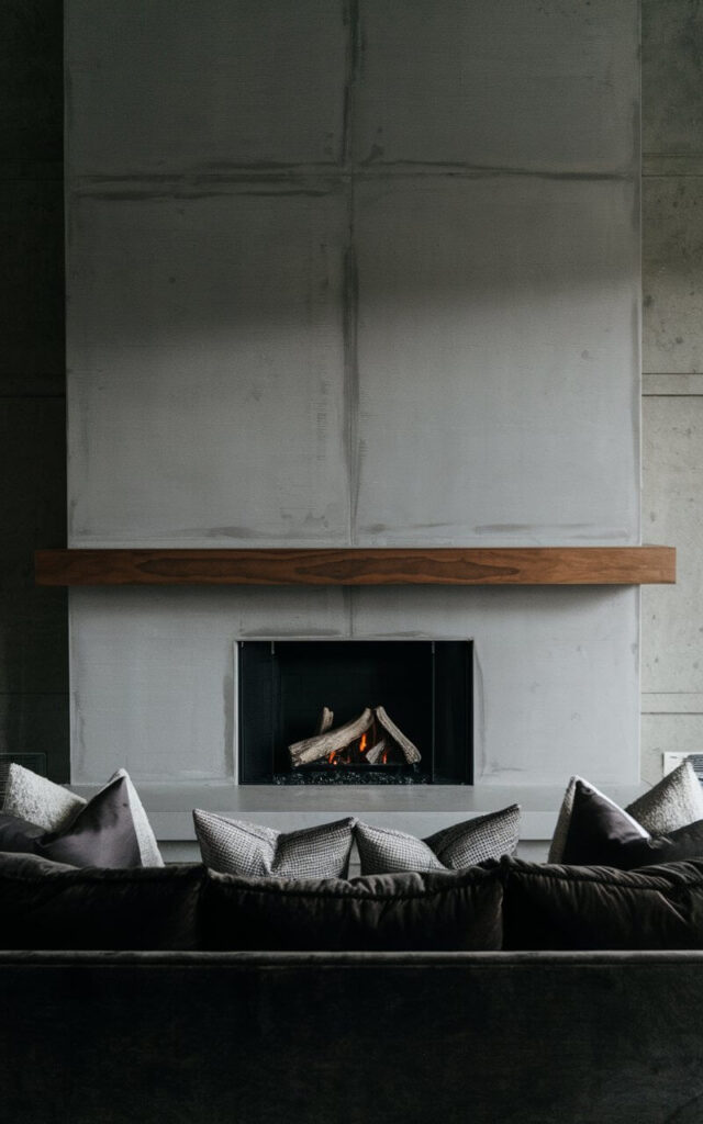 A modern, minimalist concrete fireplace makeover with clean lines and an industrial aesthetic. The smooth gray concrete surround extends seamlessly to the ceiling, giving the space a refined yet edgy look. A streamlined floating wood mantel provides a touch of warmth. Positioned in front of the fire, a very cozy sofa in a deep charcoal hue balances the space, with soft accent pillows adding texture. The room’s dim lighting enhances the moody elegance.