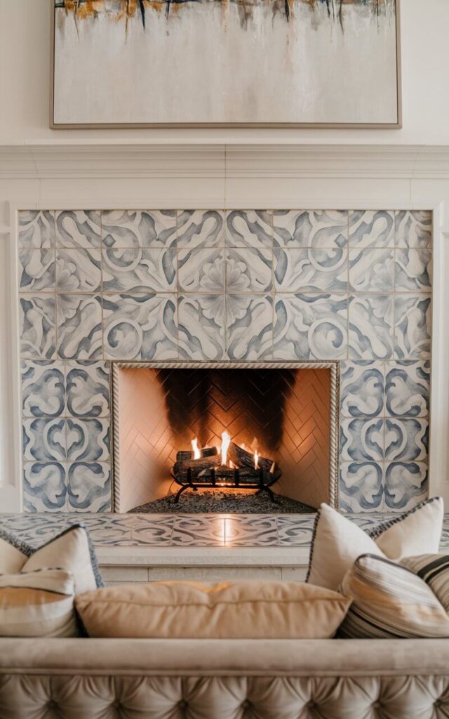 A cozy living room with a beautifully tiled hearth fireplace. The fireplace has an intricate patterned tile design in subtle blue and gray hues, with a crisp white surround. Above the mantel, there is a large abstract painting. In front of the hearth, there is a sofa with a tufted back and throw pillows. The warm firelight enhances the vibrant details of the tile.