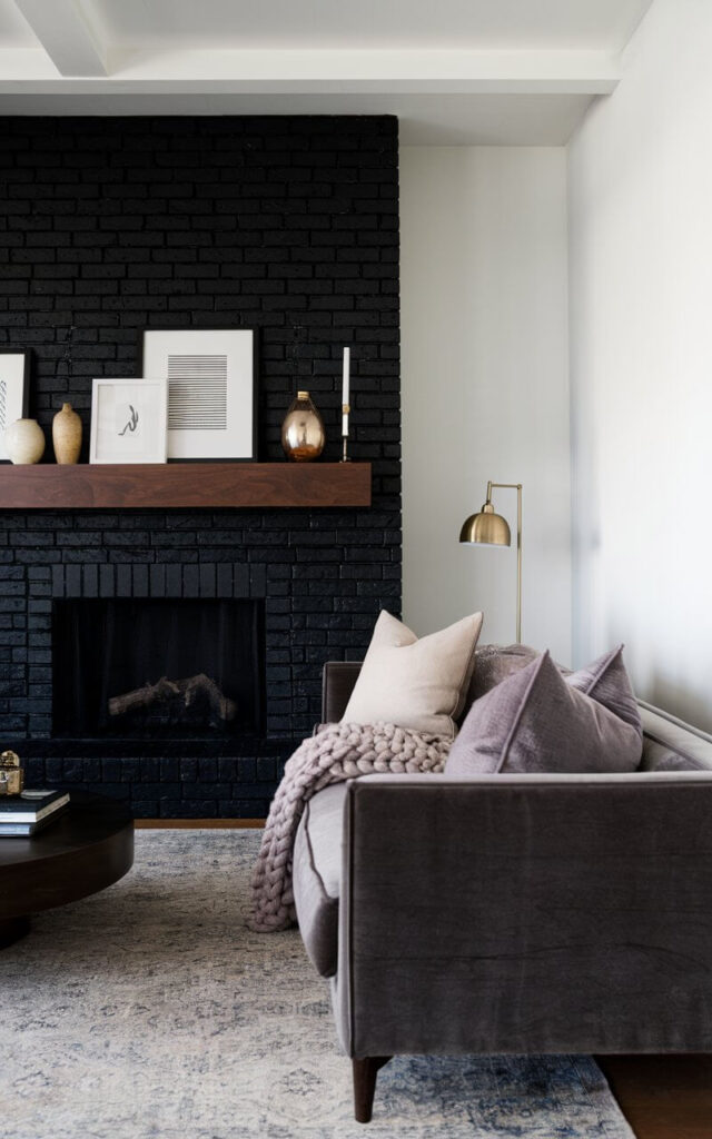 A contemporary living room with a black brick fireplace as the focal point. The deep matte black bricks contrast against the crisp white walls, creating a modern and sophisticated look. A sleek wooden mantel holds a mix of decorative vases and framed artwork. Facing the fireplace, a cozy sofa in soft gray fabric is adorned with plush throw pillows and a chunky knit blanket, enhancing the inviting ambiance. A brass floor lamp beside the sofa adds warmth, while a textured area rug grounds the space, making it feel chic yet comfortable.