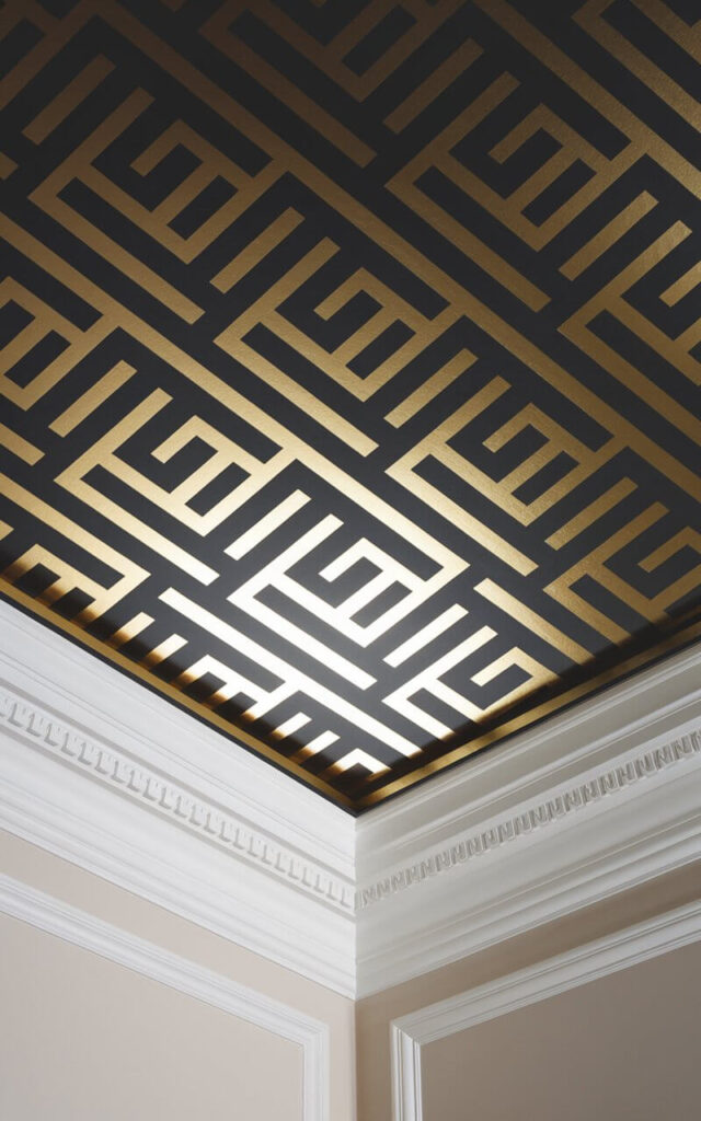 A photo of a close-up view of a ceiling wallpaper with a bold geometric pattern in black and gold. The intricate design creates a striking contrast against the smooth white crown molding, drawing the eye upward. The wallpaper’s metallic sheen reflects ambient light, adding a touch of glamour to the modern interior.