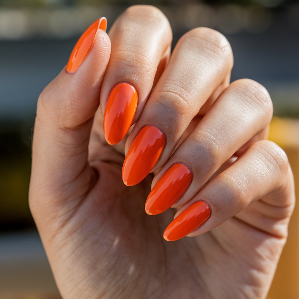 A close-up photo of a well-moisturized woman's hand displaying a vibrant orange manicure on all five fingers. The nails are almond-shaped and coated with a glossy, fiery orange polish that reflects the natural background lighting. The smooth, even application highlights the bold color, creating a striking contrast against the woman's warm-toned skin. The fingers are elegantly positioned with a relaxed curl, drawing attention to the flawless cuticles and uniform shape. Sunlight enhances the polish’s high-shine effect, making the vivid hue pop. A blurred outdoor setting in the background adds depth, subtly complementing the fiery orange tones.