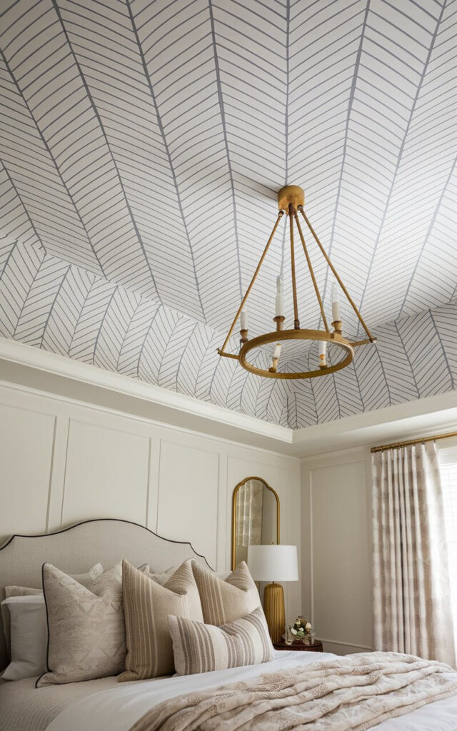 A close-up view of a ceiling wallpaper with a refined herringbone pattern in soft gray and ivory hues. The interlocking chevron lines add a structured, sophisticated touch without overpowering the space. Below, a very cozy bed with neutral-toned bedding and plush accent pillows enhances the room’s layered textures. A brass chandelier casts a warm glow, highlighting the wallpaper’s geometric precision and giving the space an effortlessly chic aesthetic.