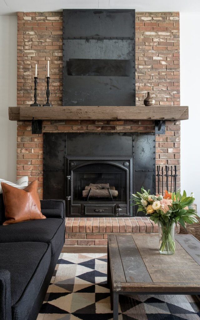 A photo of a living room with a bold brick fireplace enhanced with industrial metal accents. The fireplace features a blackened steel surround that frames the firebox, giving it a modern, edgy appeal. The exposed red brick contrasts sharply with the dark metal, creating a striking focal point. Overhead, a reclaimed wood mantel supports iron candlesticks and a minimalist vase. A very cozy sofa faces the fireplace, its deep cushions wrapped in dark charcoal fabric with a leather throw pillow adding a touch of texture. A distressed wood coffee table with a vase of fresh flowers sits atop a geometric rug, reinforcing the industrial-chic aesthetic while maintaining a welcoming and comfortable ambiance.