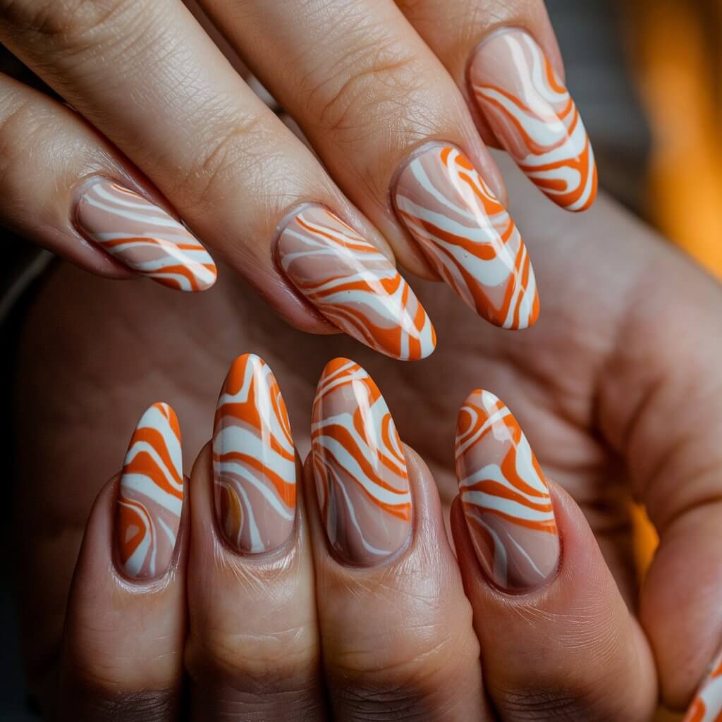 A close-up shot of a woman's hand with long, almond-shaped nails. The nails have a glossy nude base and feature intricate orange and white swirls. The orange swirls have a retro, artistic effect. The nails are well-moisturized. The background is blurred and has warm tones.