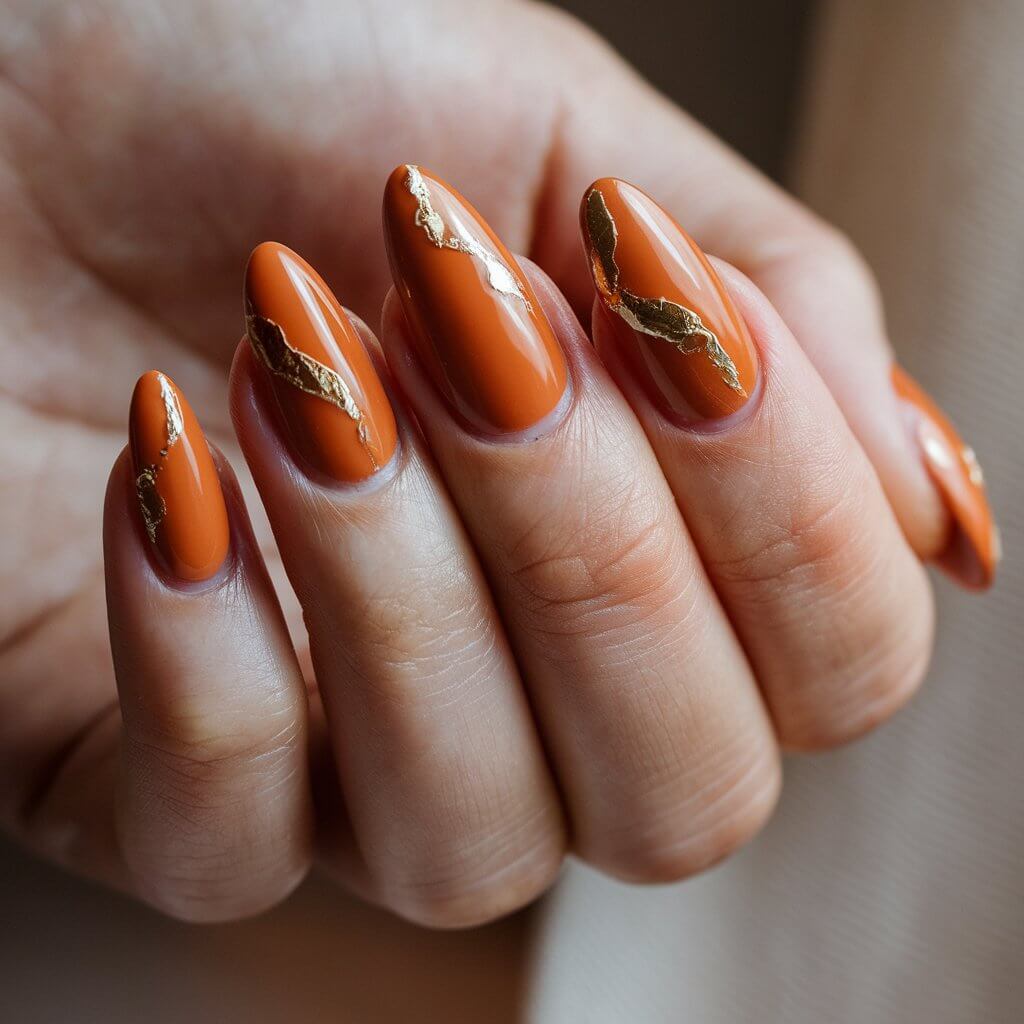 A close-up shot of a woman's hand with a well-moisturized skin. There are five fingers, each with a stunning orange manicure. The nails are almond-shaped and have a glossy burnt-orange base. There are delicate gold foil accents on the nails. The natural background lighting enhances the reflective quality of the metallic details. The fingers are positioned to allow the foil pieces to catch the light beautifully. The background is softly blurred and neutral-toned.