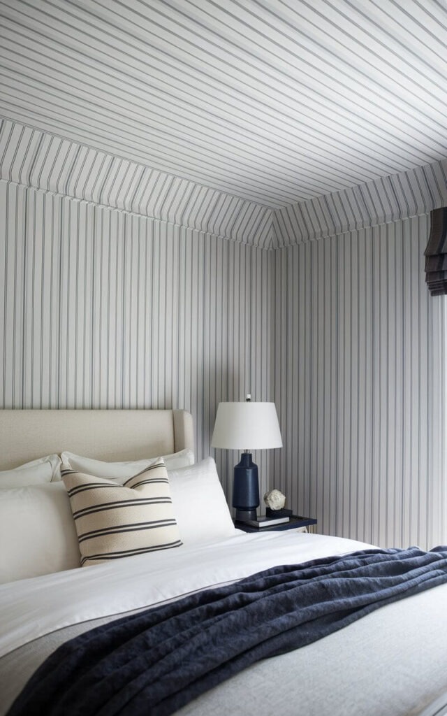 A close-up view of ceiling wallpaper featuring thin, delicate pinstripes in muted gray and white. The design adds understated elegance. Below, a very cozy bed with crisp white linens and a soft navy throw complements the wallpaper’s refined, tailored aesthetic.