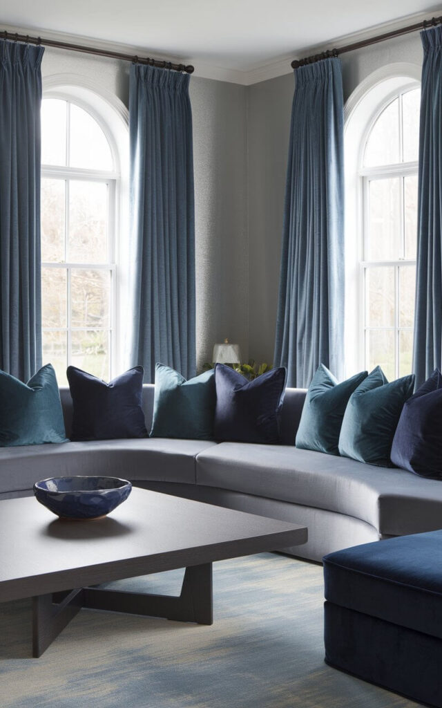A photo of a blue and grey living room with a sprawling grey sectional sofa. The sofa curves softly around a square wood coffee table, which is topped with a navy ceramic bowl. Throw pillows in all shades of blue—from powder to midnight—pepper the sectional, making it both stylish and inviting. Behind the seating area, the walls are painted a cool silver-grey, reflecting light from oversized windows dressed in steel blue curtains. A navy ottoman rests nearby, doubling as a tray table or extra seat.