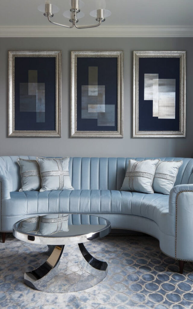 A photo of a blue and grey living room with hints of metallic. There is a plush light blue velvet sofa and pale grey throw pillows with silver thread. A chrome base round coffee table with a mirrored top is placed in the room. A trio of abstract prints in shades of navy and pewter, framed in metallic-finished wood, is mounted on the wall. A silvery blue patterned rug is placed on the floor. The room has a brushed nickel chandelier hanging overhead.