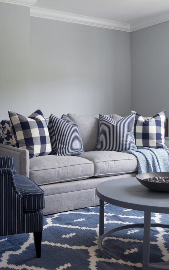 A photo of a blue and grey living room with a mix of patterns, centered around a cozy grey sofa. The sofa has throw pillows in stripes, checks, and subtle florals in navy and powder blue. The walls are painted a neutral soft grey. A blue-and-white ikat patterned rug covers the hardwood floor. A navy accent chair with a subtle pinstripe pattern is placed near the sofa. The space contains a round wooden coffee table with a grey pebbles bowl. Layered textiles and varied motifs fill the space with a sense of creativity and warmth.