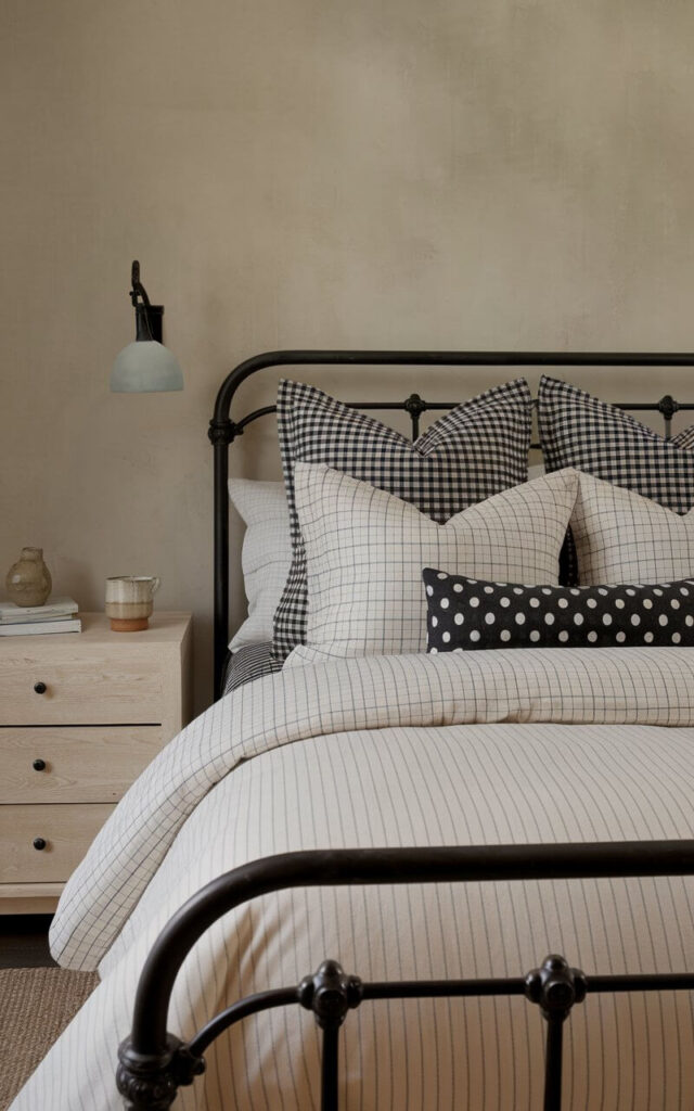 A photo of a cozy black and beige bedroom with subtle patterns. The room has a dark metal bedframe, a beige wall, and a light wood nightstand with black pulls. The bed is dressed in well-decorated bedding with layers of stripes, grids, and solids in a harmonious palette. The duvet has a beige-and-white pinstripe, and there are black plaid euro shams and a polka dot lumbar pillow. The nightstand holds a ceramic cup.