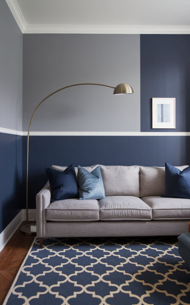 A photo of a stylish blue and grey living room with a creative two-tone wall treatment. The bottom half of the wall is painted a deep navy, while the upper half is a warm dove grey, divided by a crisp white chair rail. In front sits a cozy sofa in heather grey, its deep seat piled with cushions in varying shades of blue. A navy and ivory area rug grounds the room, and a slim gold floor lamp arches over the space. On the wall, minimalist artwork bridges the color break, pulling the eye upward. This room feels intentional and thoughtfully layered—perfect for those who love visual structure without sacrificing warmth.