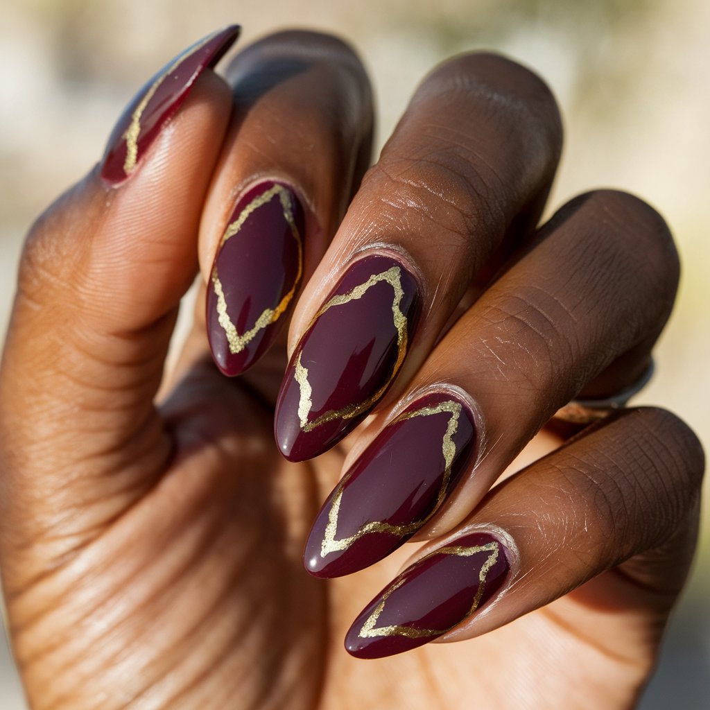 A close-up shot of a well-moisturized oily black woman's single hand, highlighting five almond-shaped nails with rich burgundy polish and negative space designs edged in ultra-thin gold striping. The nails have a high-gloss finish that glistens in the natural bright outdoor blurry background. The hand is in a relaxed pose, elegantly displaying the sharp contrast of deep red and shining gold. These Thanksgiving nails are bold, modern, and luxuriously festive.