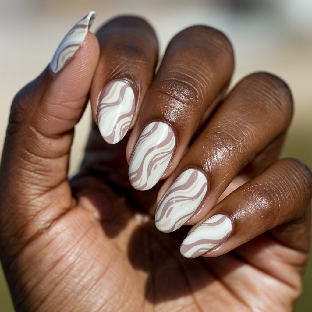 A close-up of a well-moisturized oily black woman's single hand highlights five fingers featuring an abstract marble design in creamy white and light taupe, mimicking the swirl of mashed potatoes and gravy. The nails are long and oval-shaped, with a glossy finish that reflects the natural bright outdoor blurry background. The hand is held in a relaxed pose, allowing the subtle, edible-inspired pattern to shine. These thanksgiving nails are tasteful with a clever nod to comfort food.