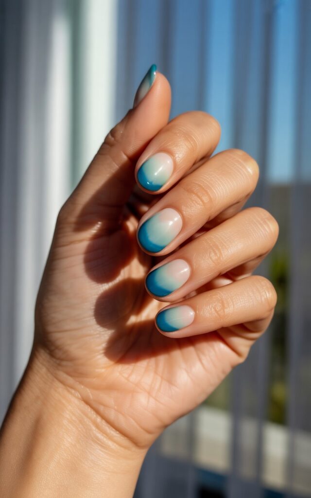 A photograph of a woman's hand gently cupped, the focus solely on its smooth, well-moisturized skin and striking nail design. Each of the five fingers features an ombré design seamlessly blending from a soft seafoam green at the base to a deep ocean blue at the tips, reflecting the light. The hand rests against a blurred background of sheer white curtains diffusing bright sunlight, emphasizing the glossy finish and layered tones of the nails. Soft, natural lighting illuminates the hand, evoking a feeling of warmth and tranquility, as if basking in a gentle seaside breeze.