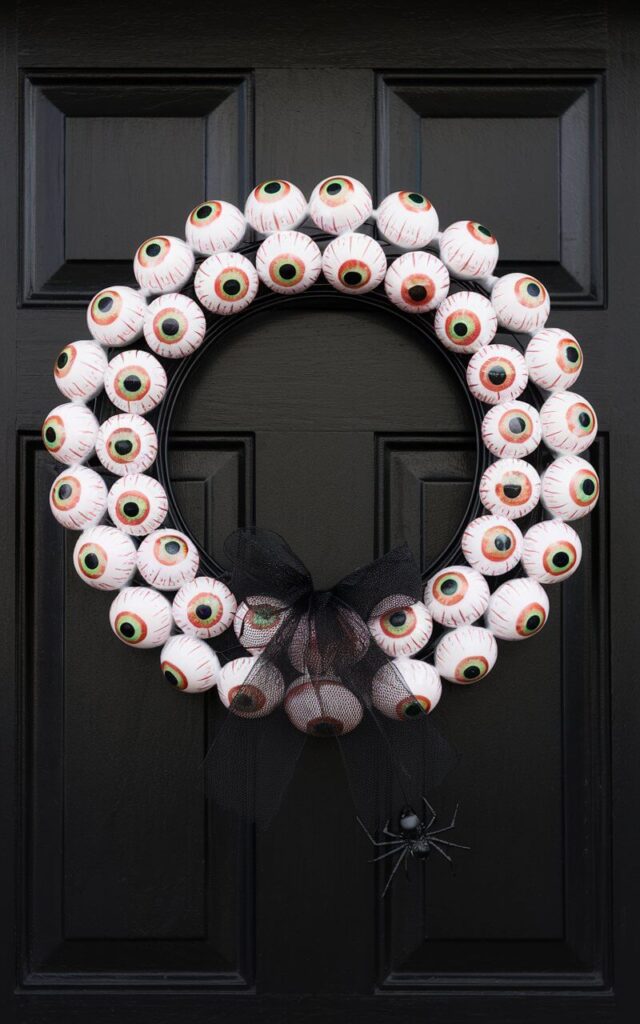 A Halloween door hanger shaped like a circular wreath of eyeballs adorns a black-painted door. The wreath is made of glossy resin eyeballs, each with hand-painted irises and bloodshot veining. The eyeballs are clustered tightly around a slim black wire frame, creating a clean circular form. A black mesh ribbon and a single dangling faux spider accent the bottom. The monochromatic base allows the vivid eyes to stand out dramatically, giving the hanger a surreal, watchful presence that's both eerie and captivating.