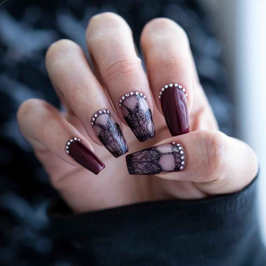 A photo of a woman's hand with five coffin-shaped nails in a deep burgundy colour. The nails have a gothic lace design in black, with tiny rhinestones at the edges. The background is blurry and bright, contrasting with the dark tones of the nails and the hand. The hand is in a relaxed pose, with the fingers slightly curled inward.