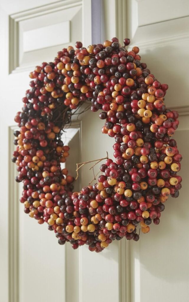 A photograph of a meticulously crafted fall wreath hanging on a pristine white door. The wreath is densely packed with berry clusters in a rich palette of cranberry, burnt orange, and deep plum; each berry displaying a subtle sheen and faint speckles. Tiny twigs weave through the clusters, adding depth and contrasting textures, while pinpoint highlights accentuate the berry skins. Soft, diffused sunlight illuminates the wreath and door, casting gentle shadows and creating a warm, inviting atmosphere.