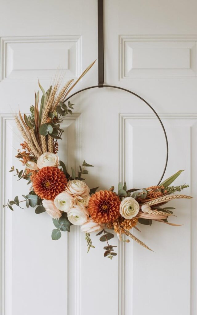 A photograph showcasing a meticulously crafted Fall wreath suspended against a stark white door. The wreath features a sleek matte black metal hoop partially wrapped with a lush arrangement of rust-colored dahlias, ivory ranunculus, eucalyptus sprigs, and wispy wheat strands, all bound with copper wire and delicate floral tape. The unadorned upper arc of the hoop creates a deliberate negative space, highlighting the floral density and emphasizing the wreath’s clean, contemporary design. Soft, diffused daylight illuminates the scene, casting gentle shadows that accentuate the wreath's texture and elegant form.