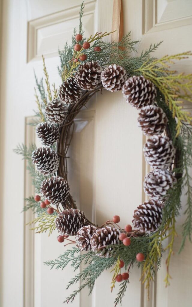 A photograph of a beautifully crafted Fall wreath hanging on a pristine white door. The wreath features an intricate arrangement of pinecones in varying sizes, their scales displaying a subtle matte finish, interwoven with fine evergreen branches exhibiting shades of deep green and muted sage. Small, muted red faux berries are nestled at the base of a few cones, providing gentle contrast to the organic textures of the grapevine core that peeks through in places. Soft, natural light illuminates the scene, highlighting the wreath's layered precision and balanced rustic-seasonal structure.