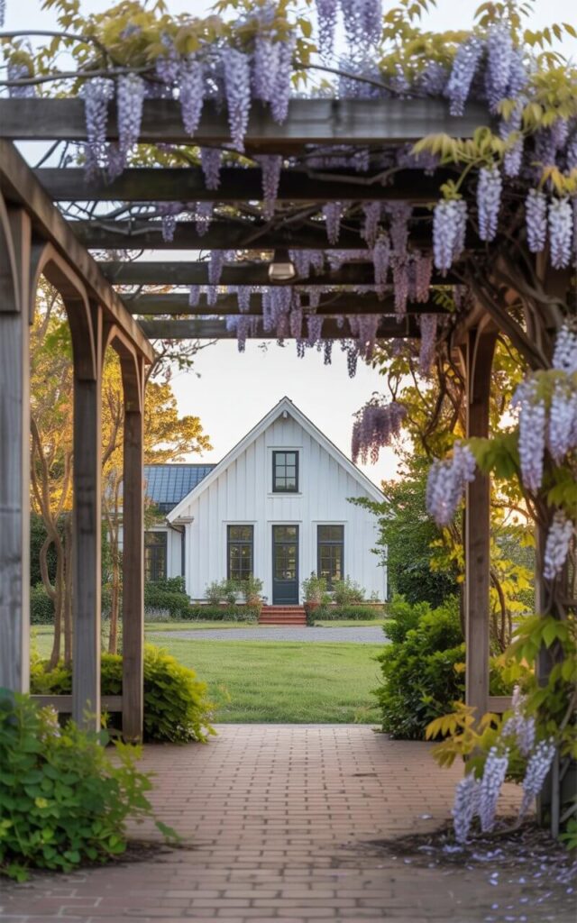 A photograph of a beautifully designed farmhouse home framed by a charming wooden pergola. The farmhouse presents a crisp white exterior contrasted by sleek black-framed windows and warm cedar trim, standing proudly against a backdrop of lush greenery.  The pergola, draped with cascading wisteria blossoms in shades of lavender and violet, creates a romantic entrance to a brick patio partially shaded by the pergola’s beams. Soft golden light filters through the wisteria canopy, illuminating the farmhouse and creating a tranquil, inviting atmosphere.