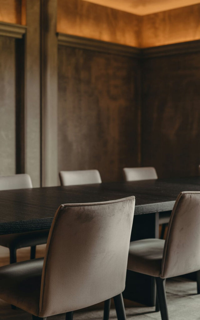 A photograph of an elegant dining room interior showcasing rich textures and matte finishes in a sophisticated minimalist design. The centerpiece is a sleek matte black wooden dining table surrounded by six minimalist chairs upholstered in soft stone gray fabric, their surfaces appearing velvety and non-reflective. The walls feature a textured plaster finish in warm charcoal tones, while subtle architectural details like built-in shelving and crown molding add depth without ostentation. Carefully positioned warm ambient lighting grazes each surface at low angles, creating gentle shadows that emphasize the tactile quality of materials while maintaining an atmosphere of quiet luxury and understated elegance.