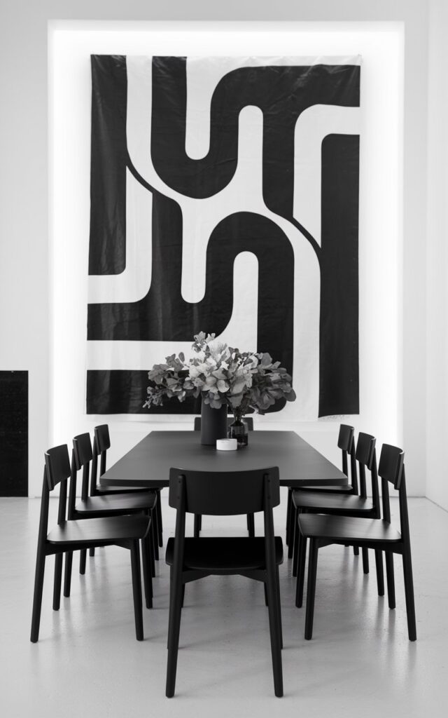 A stylish black and white dining room with a large black-and-white artwork on a bright wall. There is a plain table in the center, surrounded by minimalist black dining chairs. The artwork adds personality, depth, and visual rhythm to the room. The monochrome palette creates harmony throughout the space, while the art adds visual rhythm. The room feels curated yet understated, blending creativity with minimalist sophistication.