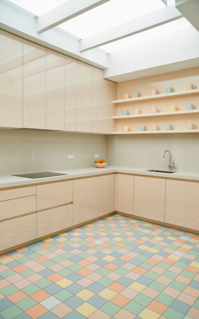 A photograph of a bright, minimalist kitchen featuring a stunning multi-color pastel checkered floor with soft mint green, baby blue, pale peach, and butter-yellow squares arranged in a playful geometric pattern. The pure white satin-finish cabinetry reflects the gentle hues of the checkered tiles, while minimalist open shelving displays coordinated pastel mugs that add charm without clutter. Clean white quartz countertops remain uncluttered except for a small pastel fruit bowl, and thin chrome fixtures provide modern accents throughout the space. Natural light floods in through a wide skylight above, causing the pastel colors to glow softly against the smooth neutral surfaces, creating a cheerful yet restful atmosphere that perfectly balances playful design with minimalist sophistication.