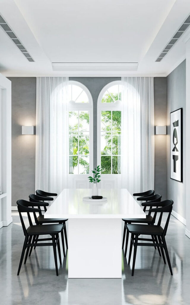 A rectangular white dining table with a smooth surface stands centered in a spacious room. Six black dining chairs with angular backs and thin legs are positioned around the table, spaced evenly apart. Large windows with sheer white curtains occupy the far wall, allowing diffused sunlight to illuminate the polished concrete floor. The room’s walls are painted a matte gray, featuring recessed lighting fixtures along the ceiling and a single framed print displaying abstract geometric shapes on one wall.