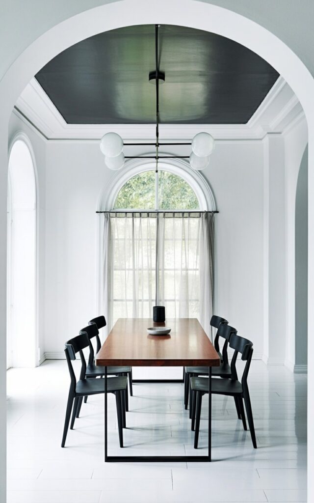 A rectangular dining table with a smooth, dark wood surface stands in the center of a room, surrounded by six simple black chairs with straight backs and slender legs. A minimalist ceiling fixture with exposed metal rods and frosted glass globes hangs directly above the table, casting a soft glow. The ceiling is painted a deep matte black, contrasting with the bright white walls that extend from floor to ceiling, showcasing subtle crown molding details. A large, arched window with sheer linen curtains occupies one wall, allowing diffused daylight to illuminate the room’s polished hardwood floor and the faint texture of the plaster walls.
