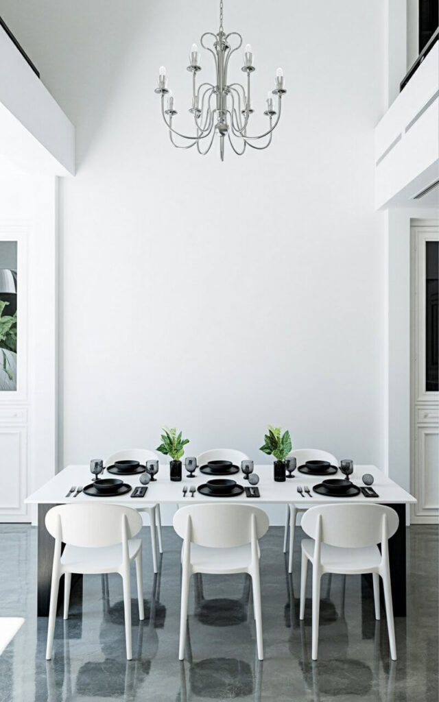 A rectangular dining table is set for a formal dinner with six place settings. Each place setting includes a black ceramic plate, a black bowl, black stainless steel cutlery, and a black glass tumbler. Six minimalist white dining chairs with curved backs are evenly spaced around the table, providing comfortable seating. The room features a polished concrete floor with a large, ornate chandelier suspended from the high ceiling above.