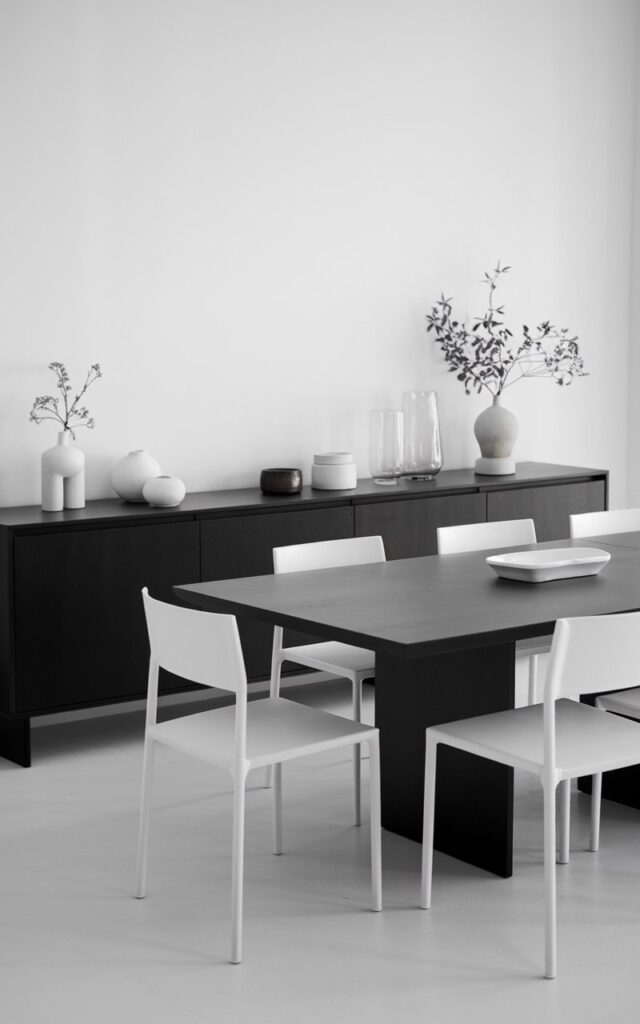 A black and white dining room with a sleek black buffet along one wall. The dining room has a plain table in the center with minimalist white dining chairs. Decorative items such as white ceramics, simple greenery, or glass pieces are placed on the buffet. The overall look of the room is clean and minimalist with the buffet providing a refined sense of structure.