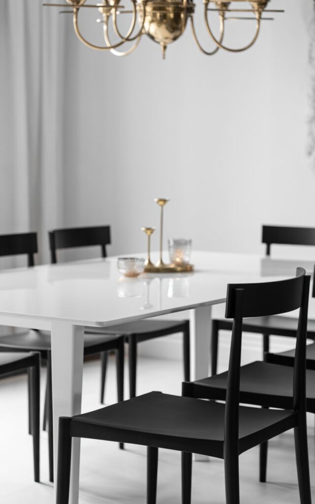 A sophisticated black and white dining room photograph showcasing elegant monochrome interior design with luxurious metallic accents. The centerpiece is a pristine white dining table surrounded by sleek black chairs featuring thin, geometric frames that create striking silhouettes against the neutral backdrop. Above the table, an ornate brass or gold chandelier casts warm, diffused light across the space, while delicate metallic candleholders on the table surface add subtle gleaming highlights. The room maintains a refined minimalist aesthetic with crisp contrast between the black and white elements, while the strategic metallic touches create depth and sophisticated glamour throughout the space.
