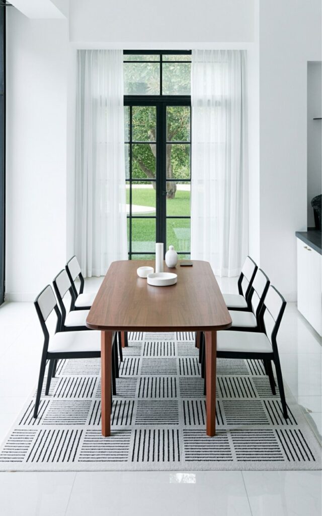 A rectangular dining table with a smooth, dark wood surface stands centered on a patterned rug. Six minimalist white chairs with straight backs and thin legs are evenly spaced around the table. The rug features a geometric pattern composed of intersecting black and white lines, creating a visual contrast against the white chairs and dark table. A large window with sheer white curtains fills one wall, allowing soft daylight to illuminate the room and revealing a glimpse of a manicured lawn outside.