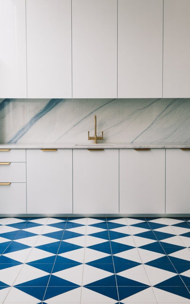 A photograph of a minimalist kitchen featuring a striking navy-and-white checkered floor with deep, saturated navy tiles contrasting against bright white tiles in a crisp geometric pattern. The sleek white cabinetry displays clean, uninterrupted lines that emphasize the minimalist design, while brushed brass hardware and faucet add warm metallic accents throughout the space. A elegant marble backsplash with subtle blue veining runs along the wall, perfectly echoing the floor's color palette and creating visual harmony. Soft, natural light floods the kitchen, highlighting the refined contrast between the bold checkered floor and the serene white surfaces, creating an airy yet structured atmosphere.