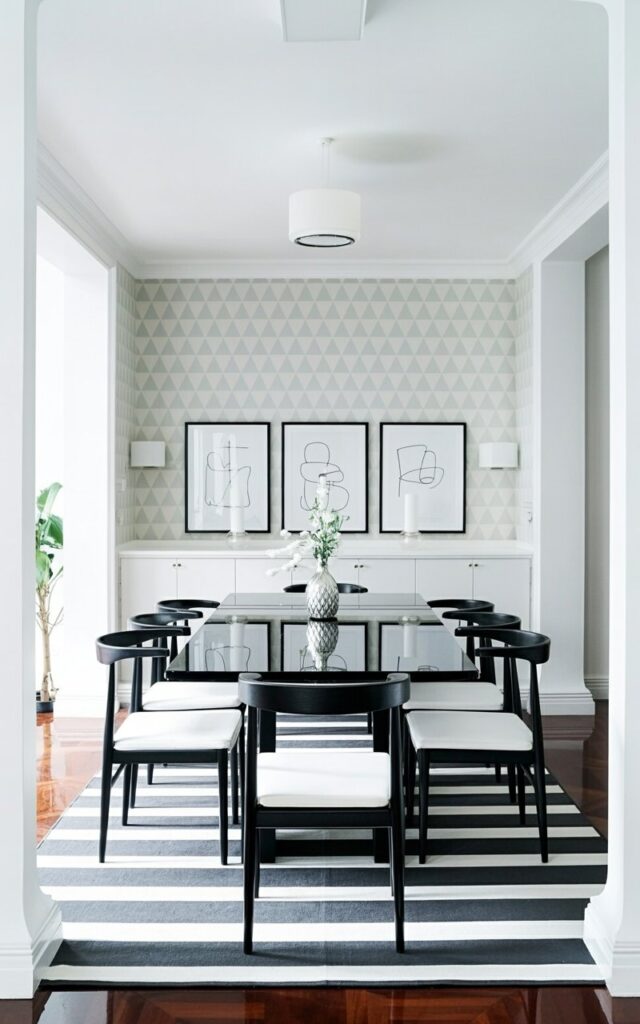 A black dining table sits centered within the room, its surface clear and reflecting the overhead lighting. Six minimalist black dining chairs with crisp white square cushions surround the table, each positioned evenly apart. The walls are covered in a subtle geometric wallpaper pattern featuring repeating triangles in varying shades of gray, complemented by three framed prints displaying abstract line art in black and white. A bold striped rug with thick horizontal lines of charcoal gray and off-white anchors the table, extending to the edges of the room and showcasing a polished hardwood floor beneath.