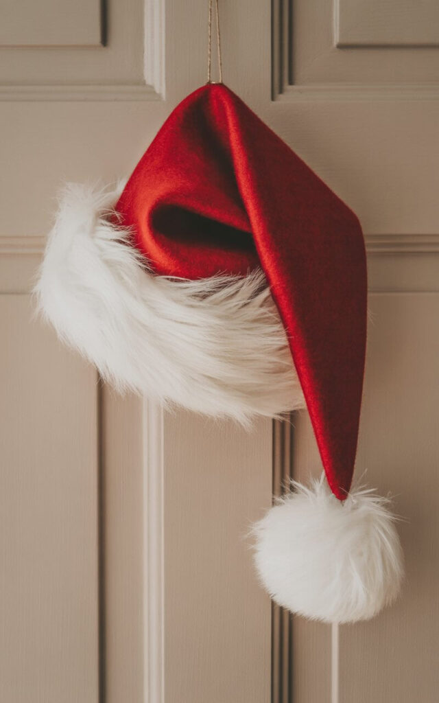 A minimalist Christmas door decoration featuring a large red felt Santa hat hanging gracefully on a smooth beige door. The hat displays rich crimson fabric with plush white faux fur trim around the brim and a generous fluffy white pom-pom at the tip, creating delightful textural contrast. The simple composition emphasizes the hat's soft curves and vibrant color against the neutral door surface, with gentle natural lighting highlighting the felt's matte finish and the fur's subtle shimmer. The clean, uncluttered aesthetic transforms this single festive element into an elegant holiday statement that radiates warmth and cheer.