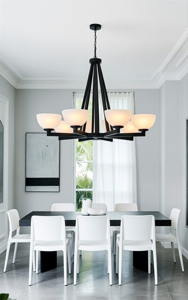A large, black chandelier with multiple arms and frosted glass shades hangs directly above a rectangular dining table. The chandelier’s metal frame features a geometric pattern, and the shades emit a soft, diffused light onto the tabletop below. Six white dining chairs with simple, straight backs are evenly spaced around the table, each chair made of molded plastic. The room has pale gray walls and a polished concrete floor, with a single, rectangular abstract painting hanging on one wall and a large window with sheer white curtains visible in the background.