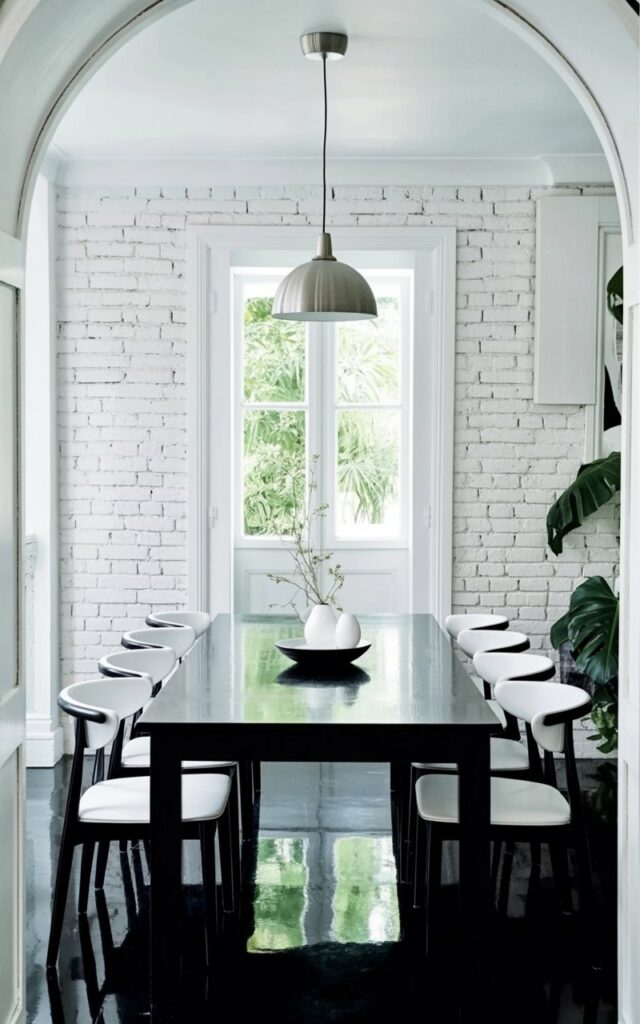 A black rectangular dining table sits centered within the room, its surface reflecting ambient light. Six minimalist white dining chairs, each with a gently curved backrest, are evenly spaced around the table's perimeter. The walls are constructed of white-painted brick, with visible mortar lines creating a subtly textured surface that extends from floor to ceiling. A simple, modern pendant light fixture with a brushed metal finish hangs directly above the table, providing focused illumination.