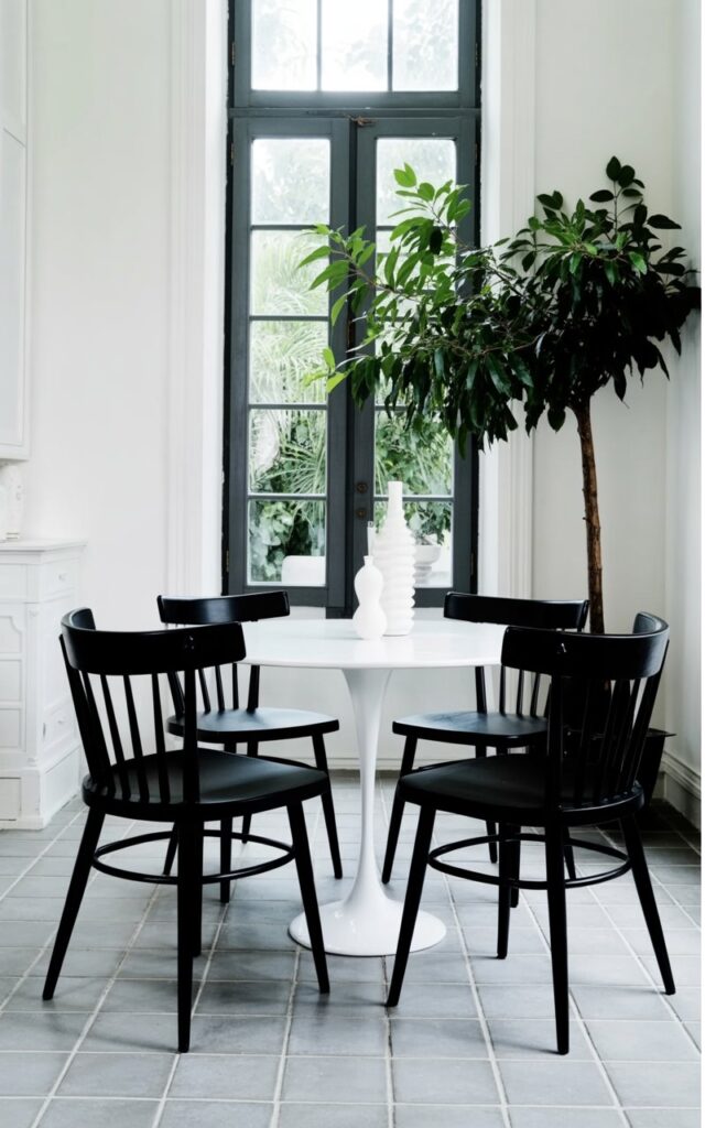 Four black dining chairs with curved backs and slender, dark metal legs encircle a rectangular white dining table. The chairs are positioned evenly around the table, with their seats angled slightly outward. A tall, narrow window with dark gray frames is visible behind the chairs, allowing natural light to illuminate the scene. The floor is covered in light gray square tiles, and a single potted ficus tree with dark green leaves sits in the corner, enhancing the room's minimalist aesthetic.