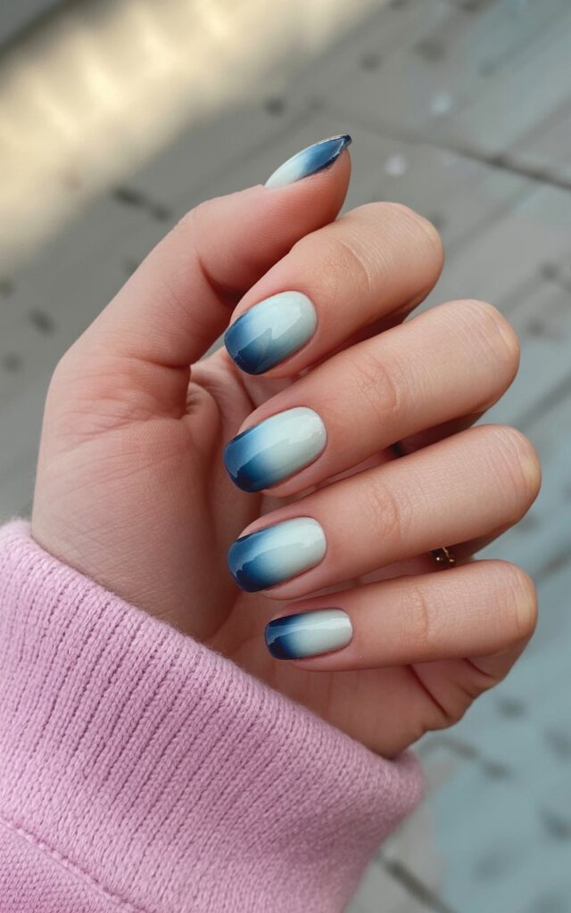 A woman's right hand rests in a relaxed position, displaying five fingers arranged with a slight curve. The nails are polished with a gradient color scheme, transitioning from a pale ice-blue hue near the cuticle to a deep midnight blue at the tips. Subtle reflections of light appear on the smooth, polished nail surfaces, highlighting the color transition. The background consists of a softly blurred field of natural sunlight, creating a gentle, out-of-focus glow behind the hand.