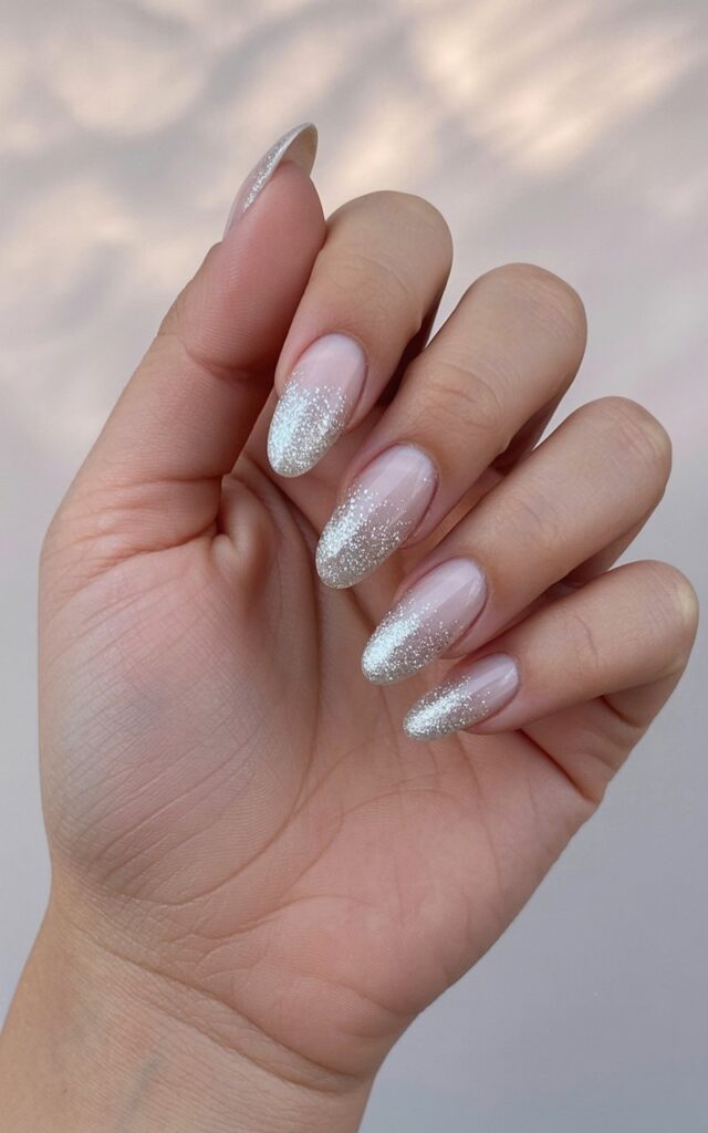 A woman's right hand is positioned palm-up with fingers slightly curved in a relaxed posture. Each of the five fingers displays long, stiletto-shaped nails coated with a base layer of shimmering silver polish, featuring concentrated glitter particles near the tips and cuticles that gradually fade outward, resembling swirling snow. The skin on the hand appears smooth and well-moisturized, reflecting soft ambient light. A gently blurred background of neutral beige tones with subtle variations in texture provides a soft, diffused backdrop.