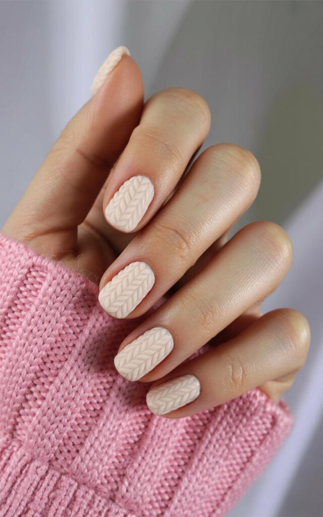 A woman's right hand rests in a relaxed position against a softly blurred background. Five fingers are slightly curved, displaying smooth, well-moisturized skin with subtle fine lines. The nails are coated in a soft beige gel polish featuring a raised cable-knit texture, resembling a cozy sweater. The hand is positioned slightly to the right of the frame, with diffused natural light gently illuminating the skin and textured nail surface.