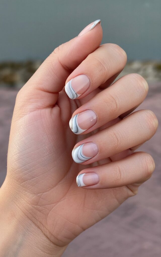 A woman's left hand rests gently on a blurred surface with the wrist slightly angled towards the viewer. Each of the five fingers is relaxed and slightly curved, displaying smooth, hydrated skin with a subtle sheen. The fingernails are nude with a frosty marble design of swirling white, silver, and pale blue across the tips, exhibiting a glossy finish and distinct marbled curves. Soft, diffused light illuminates the hand and nails against a background of muted greens and browns, creating a shallow depth of field that emphasizes the details of the hand and nails.