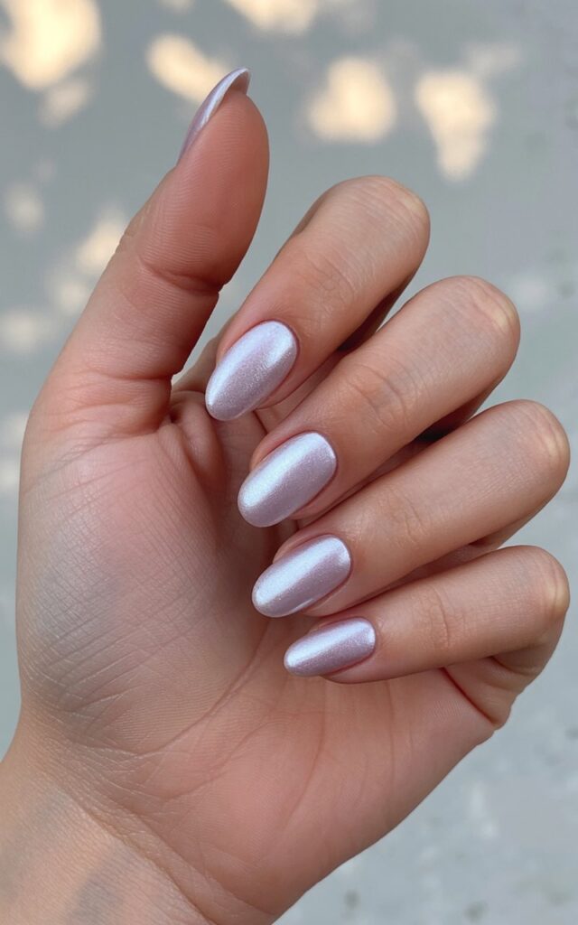 A woman's left hand rests gently on a neutral surface with the five fingers slightly curved in a relaxed posture. The hand displays smooth, well-moisturized skin with a subtle oily sheen and showcases five stiletto nails painted in muted lavender shades. A pearly, icy sheen covers the nails, creating a frosted appearance that reflects ambient light, while the cuticle area exhibits a natural pink tone. The background consists of a soft, out-of-focus field of natural light with indistinct hints of green foliage and warm brown tones.
