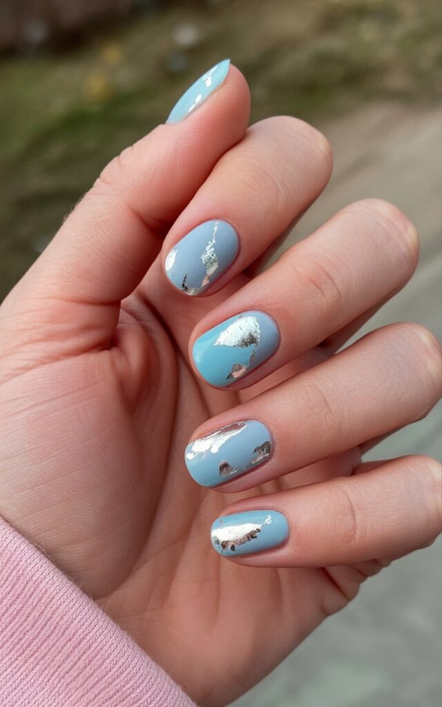 A woman's right hand rests in a relaxed position against a softly blurred natural background. Five fingers are slightly curved, showing the natural creases and lines of the skin, which appears hydrated and possesses a subtle sheen. Each nail features a base of soft icy blue with scattered pieces of silver foil adhered to the surface, creating a visual texture resembling cracked frost. Fine details of the foil reveal small imperfections and subtle reflections of light, with the hand positioned slightly off-center against a backdrop of muted greens and browns, suggesting an outdoor setting with diffused sunlight.
