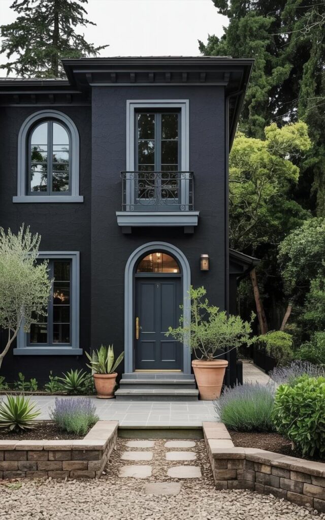 A black house exterior styled in Mediterranean architecture appears luxurious and dramatic, with smooth stucco walls painted in deep matte black. Rounded arches frame the minimalist windows, and a dark front door with a door handle anchors the entrance. Wrought iron railings and balconies add texture and charm, while stone paving leads visitors to the door. The minimalist landscaping, including potted greenery, accentuates the bold black façade. This very well designed home combines classic Mediterranean features with modern minimalist sensibilities, resulting in a striking, elegant, and confident exterior.