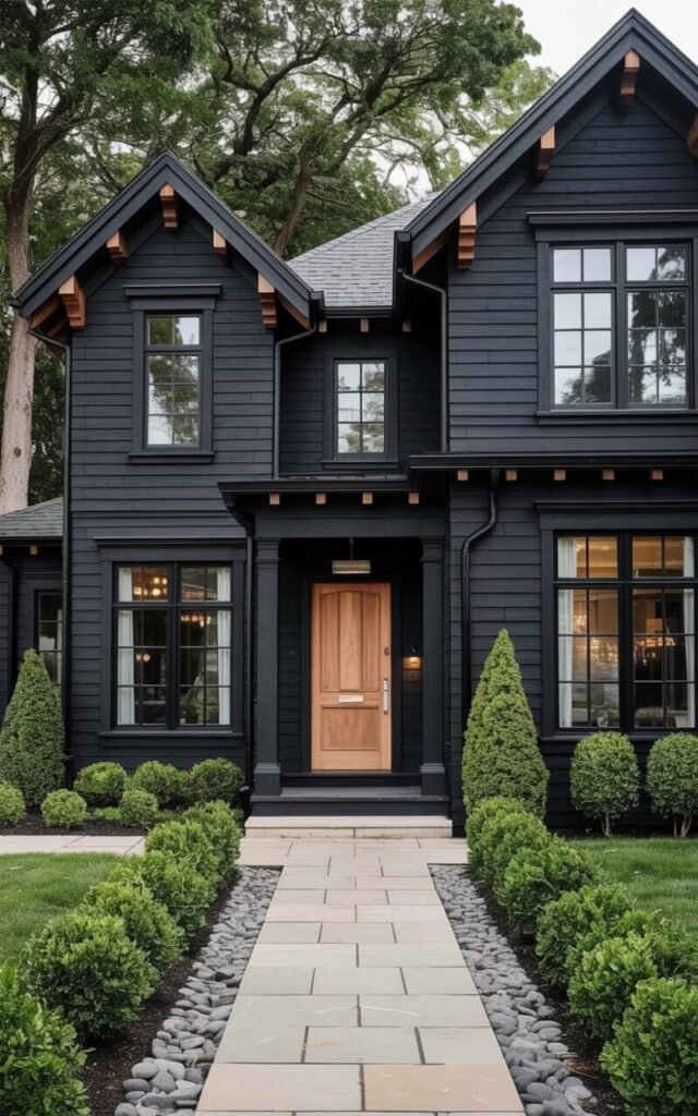 A two-story black house features a sharply angled wood-accented roofline with exposed beams along the eaves and soffits. The front door, also featuring wood accents and a brushed silver door handle, is centered on the facade, leading to a recessed entryway. Large rectangular black-framed windows are evenly spaced across the front of the house, reflecting the surrounding sky. A stone pathway leads from the driveway to the front door, bordered by neatly trimmed boxwood shrubs and smooth gray river rocks.